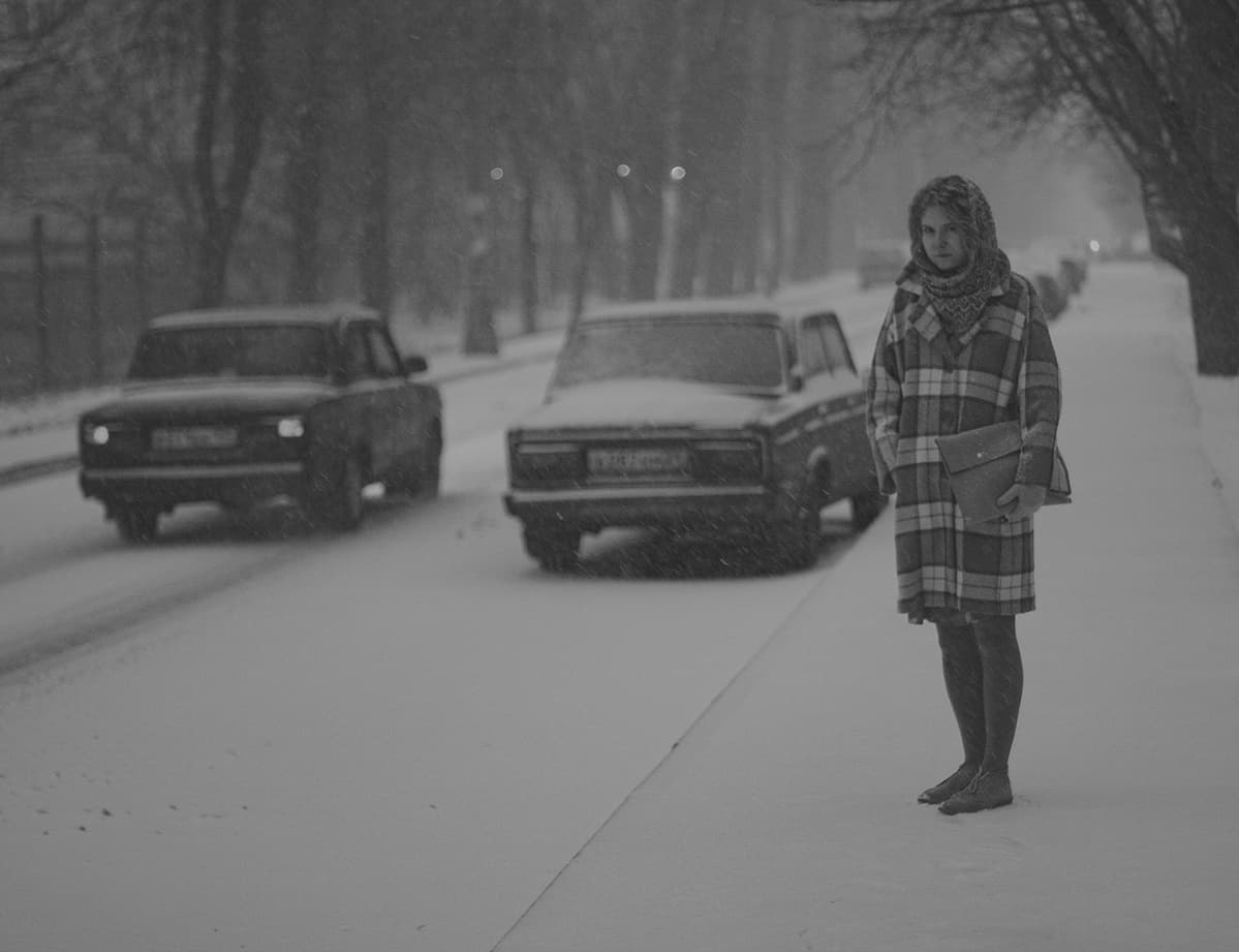 This is a young woman named Ksenia (who I am happy to call my friend) standing on the pavement of 5th Avenue in Novogireyevo, Moscow, lined with old poplars. You can see an old Lada (either model 2105 or 2107) parked nearby, with another Lada passing by.
